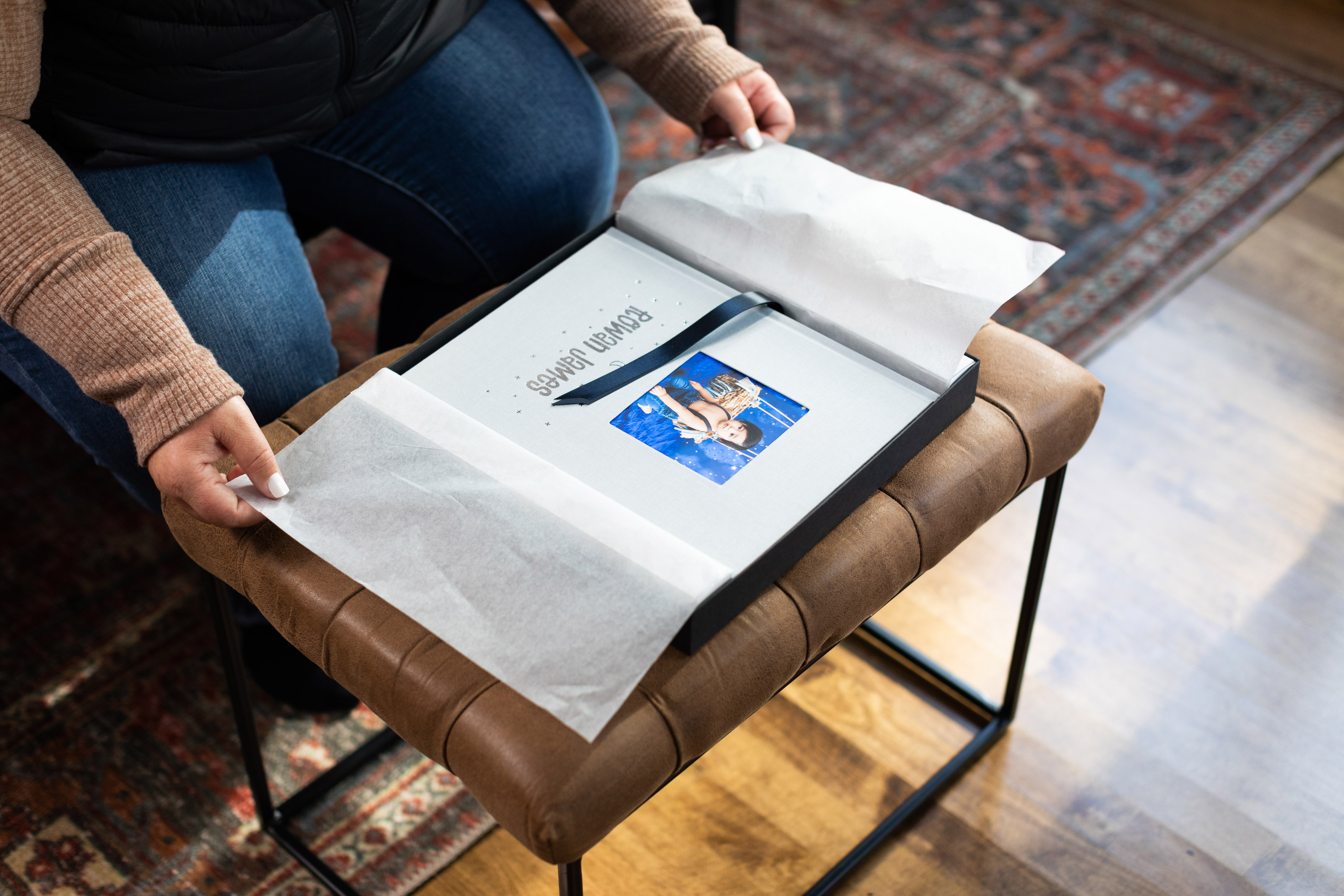 Hands open the tissue paper covering a Cameo Cover Album with a "Rowan James" custom debossed cover inside a charcoal Premium Packaging box. The box is on a small padded bench in a home.