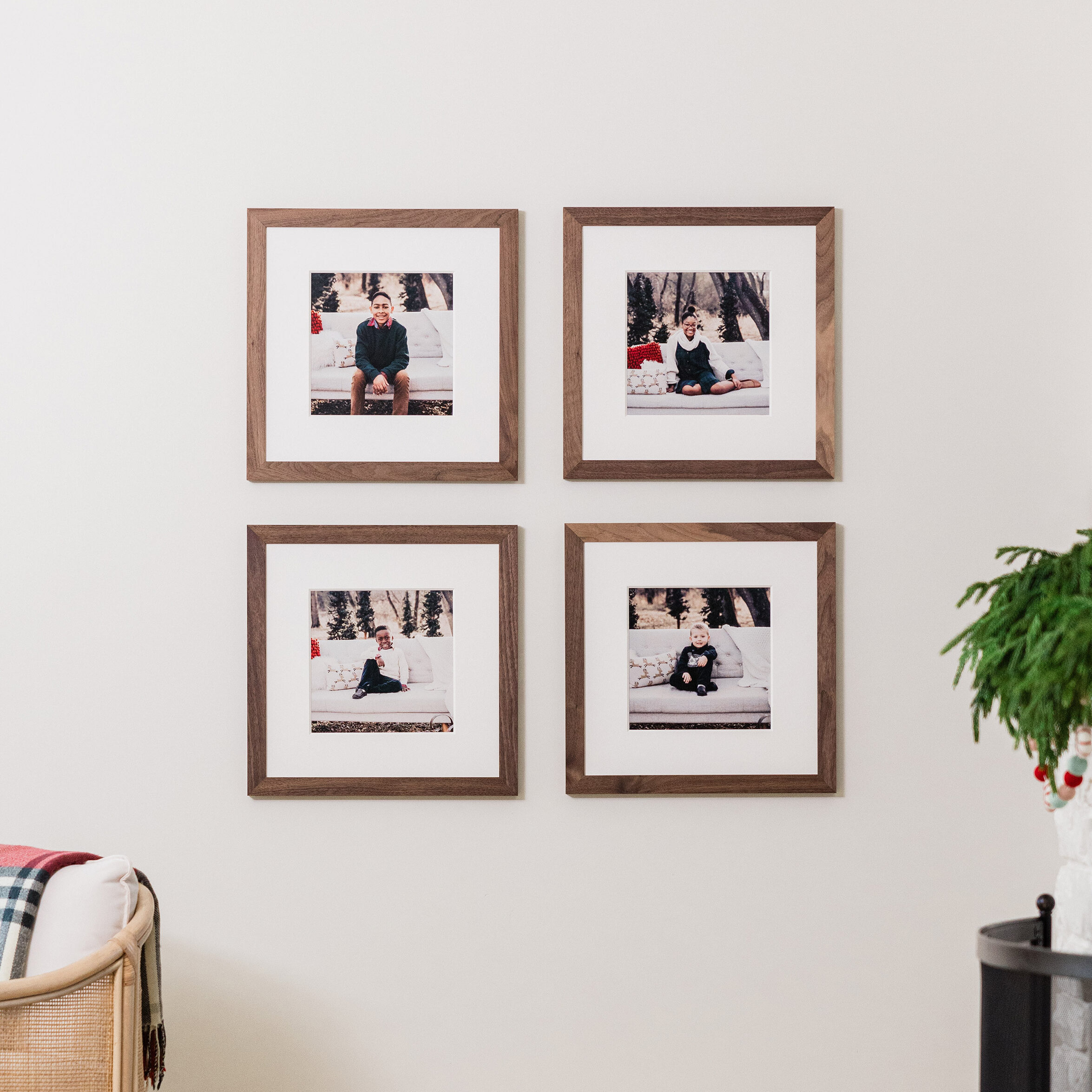 Grid wall collage of four square Walnut Woodland Framed Prints with mats hung on living room wall with fireplace and holiday garlands, featuring holiday-themed portraits of the family's children