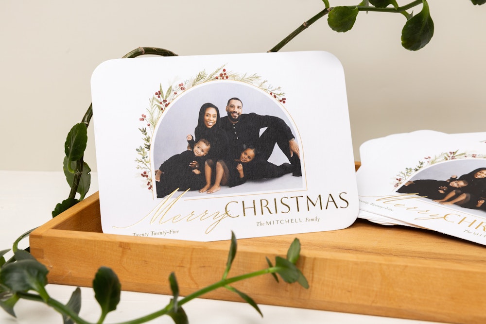 Professional family portraits in a studio printed on a high-quality holiday card featuring drawn winter greenery accents framing the image and "Merry Christmas" message foil-pressed in gold. Card is standing up in a wood tray on a white table by a plant.