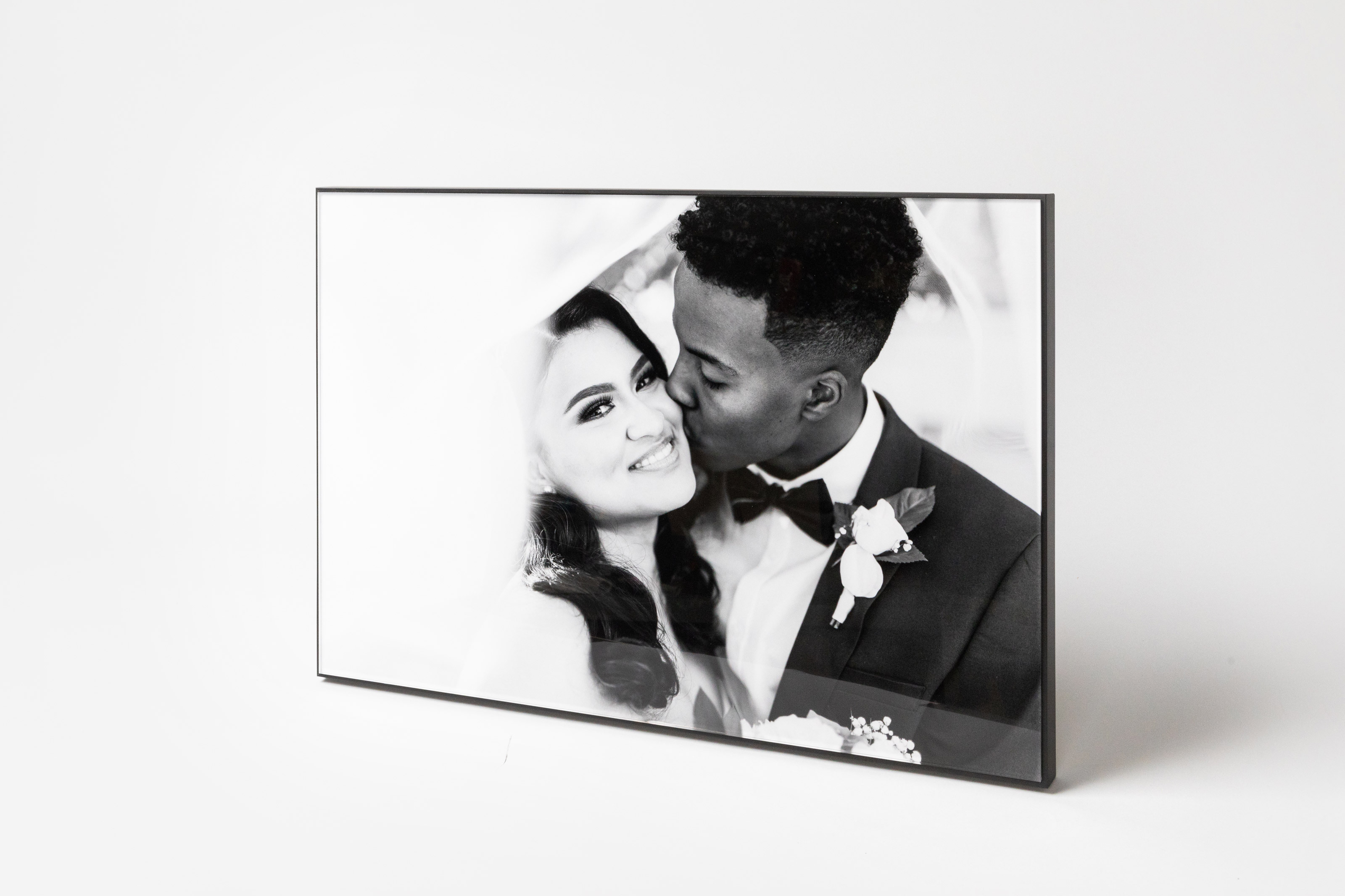 Wedding portrait of groom kissing bride's cheek as veil floats around them, displayed in a Black Framed Acrylic Print standing on a soft gray background