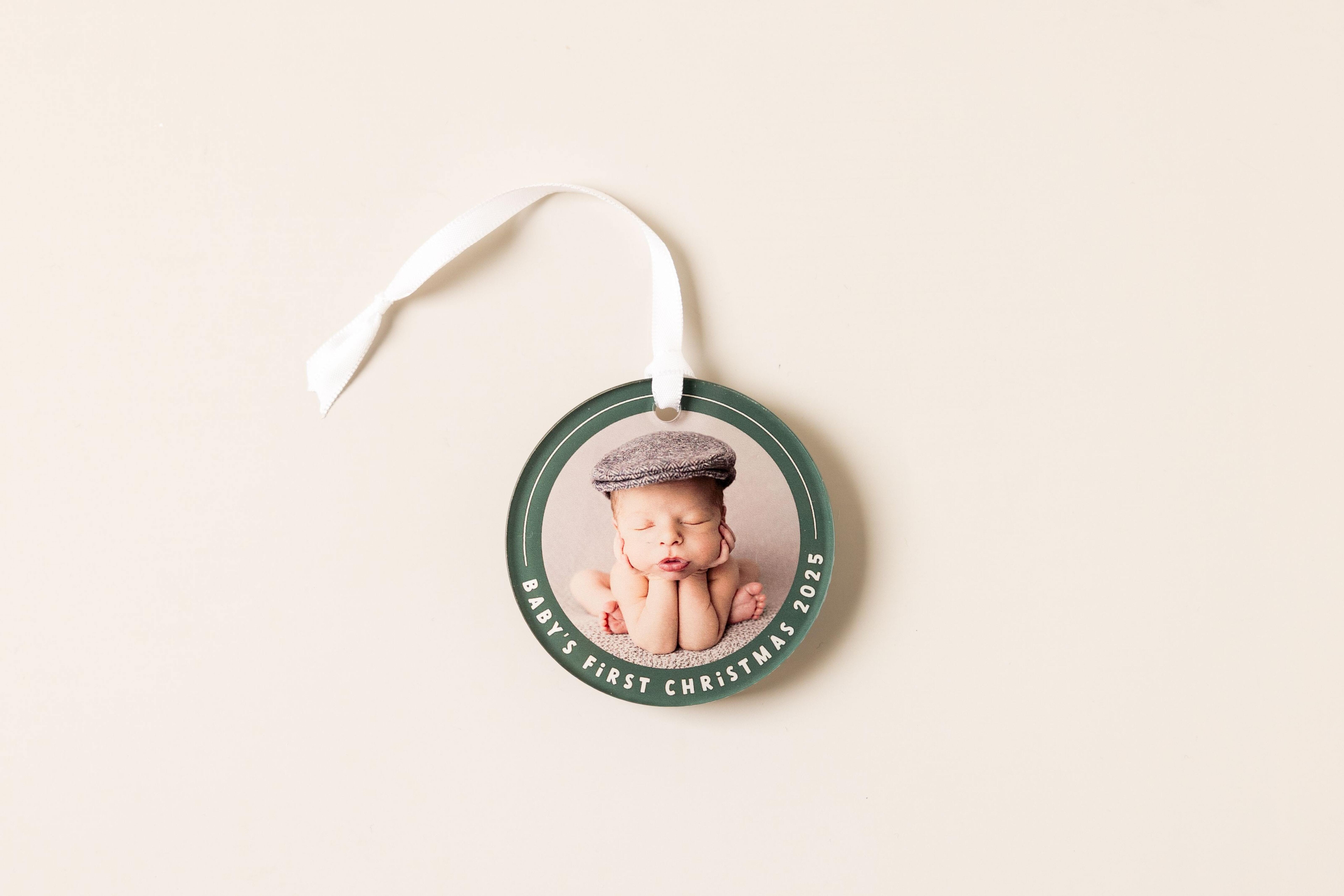 Acrylic ornament with a sleeping newborn in a knit hat, printed with "Baby's First Christmas" around a green border and hung on a white satin ribbon