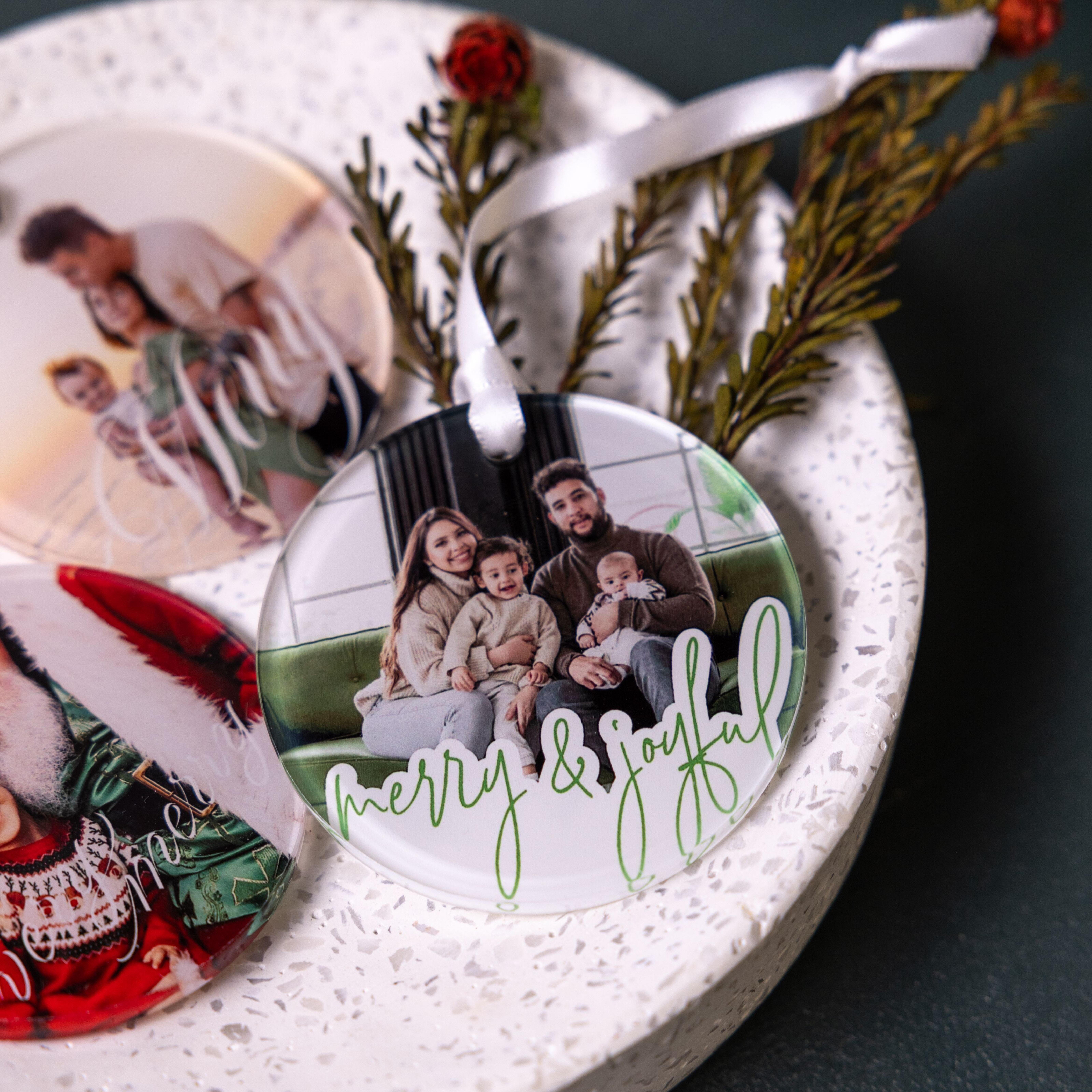 Acrylic photo ornaments with family portraits and holiday text, styled with greenery, red flowers, and a white ribbon