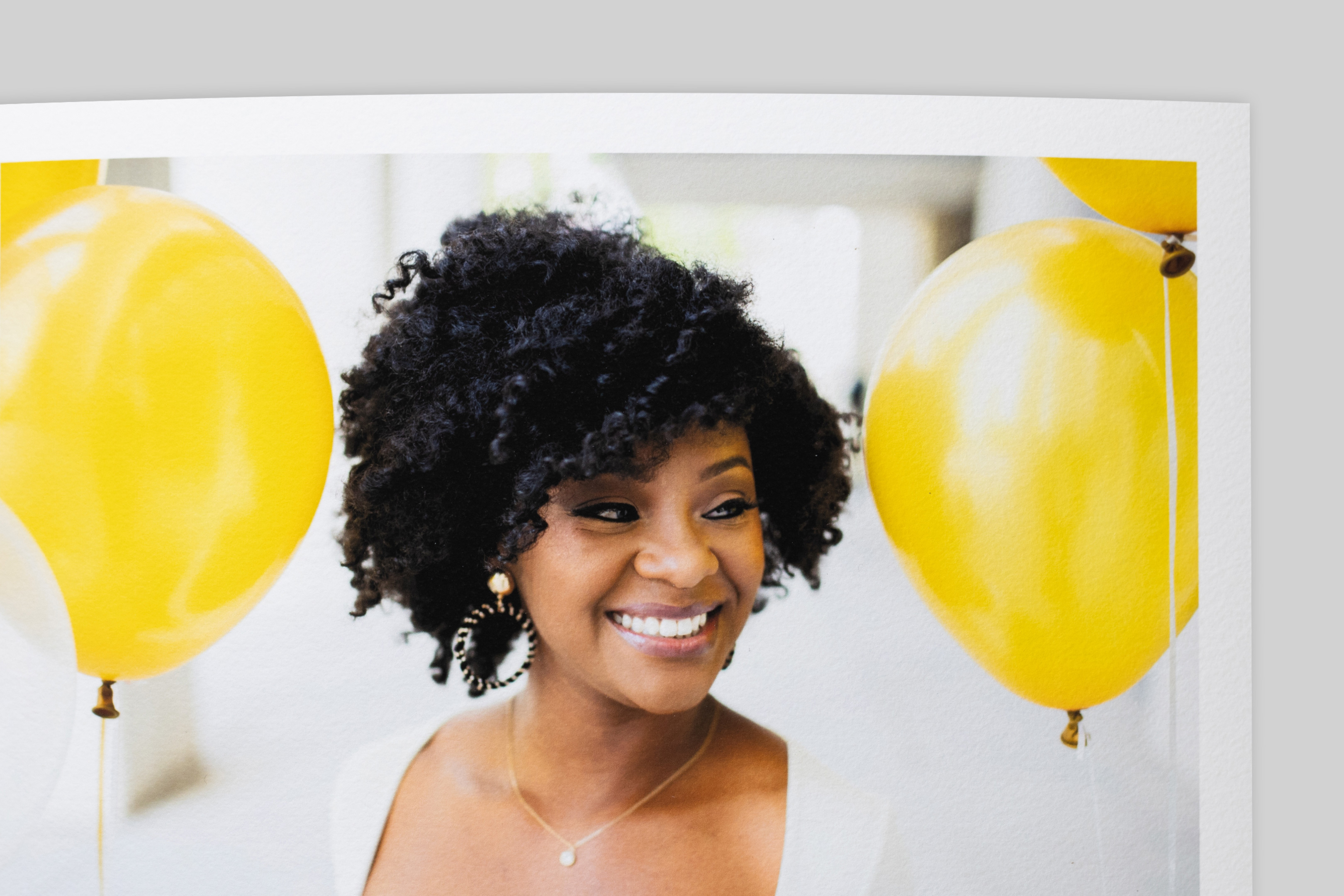 Corner of Aquarelle Rag Fine Art Print with border, featuring a portrait of a smiling woman with curly hair amid bright yellow balloons. Slight texture visible on paper surface.