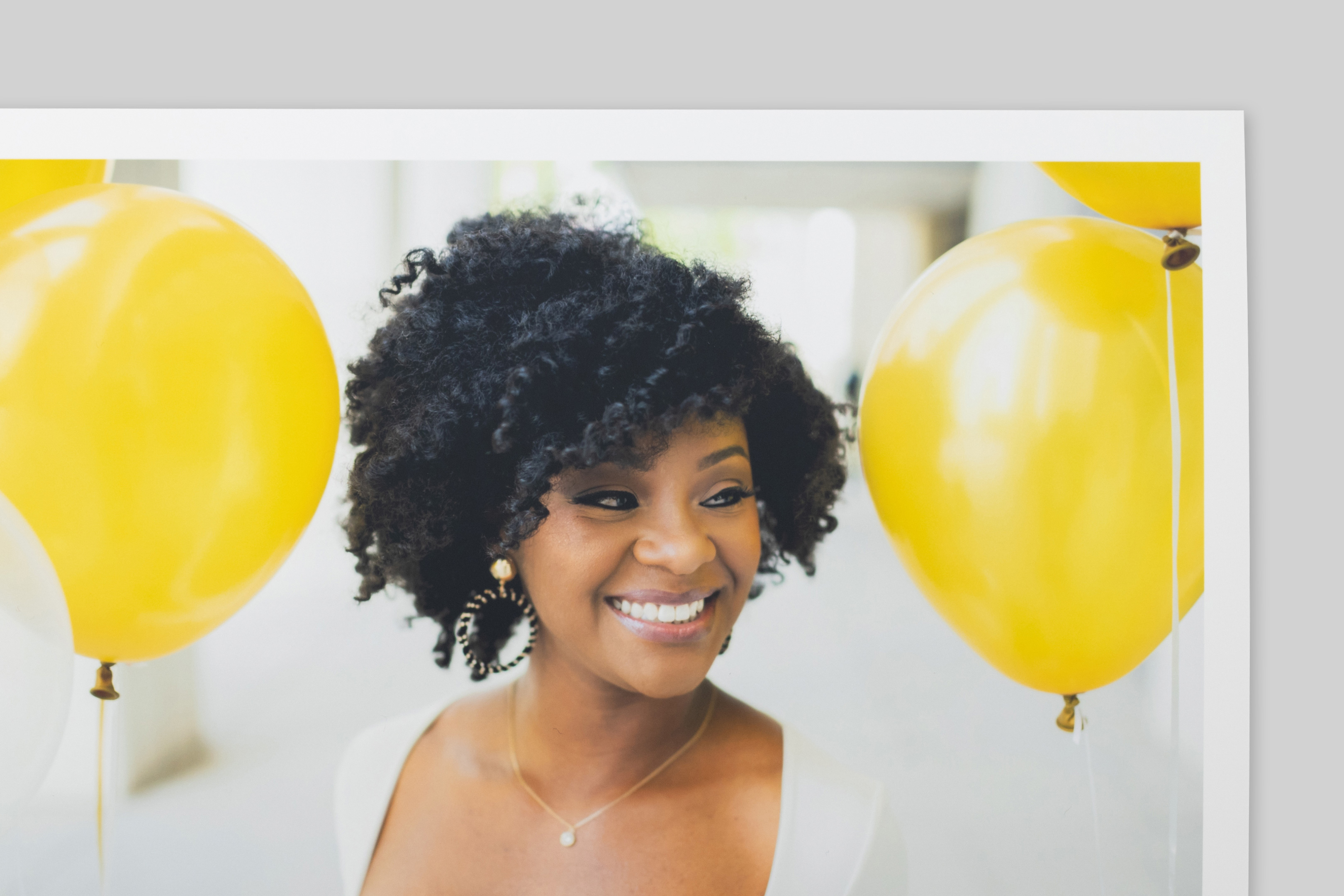 Corner of Deep Matte Photo Print with border, featuring a portrait of a smiling woman with curly hair amid bright yellow balloons. No texture or sheen visible on paper surface.