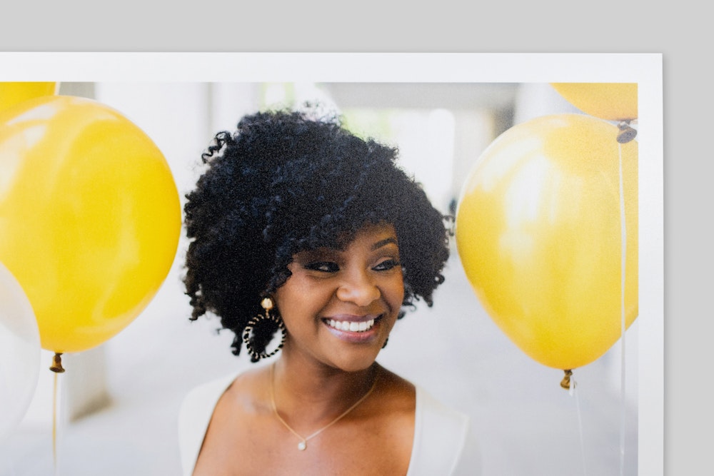 Corner of Lustre Photo Print with border, featuring a portrait of a smiling woman with curly hair amid bright yellow balloons. Very subtle sheen and texture visible on paper surface.