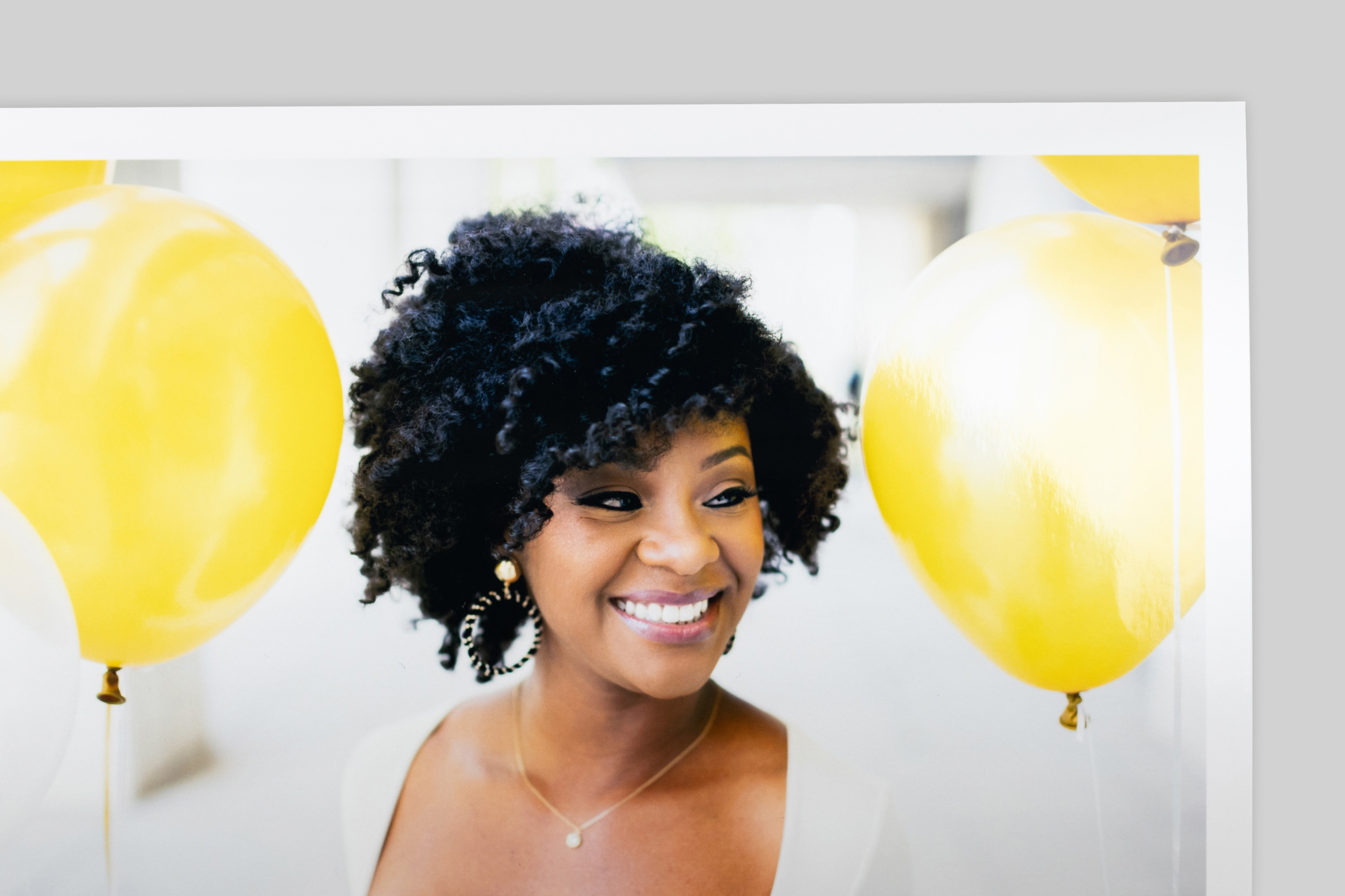 Corner of Pearl Photo Print with border, featuring a portrait of a smiling woman with curly hair amid bright yellow balloons. Strong reflection and glossy finish visible on paper surface.