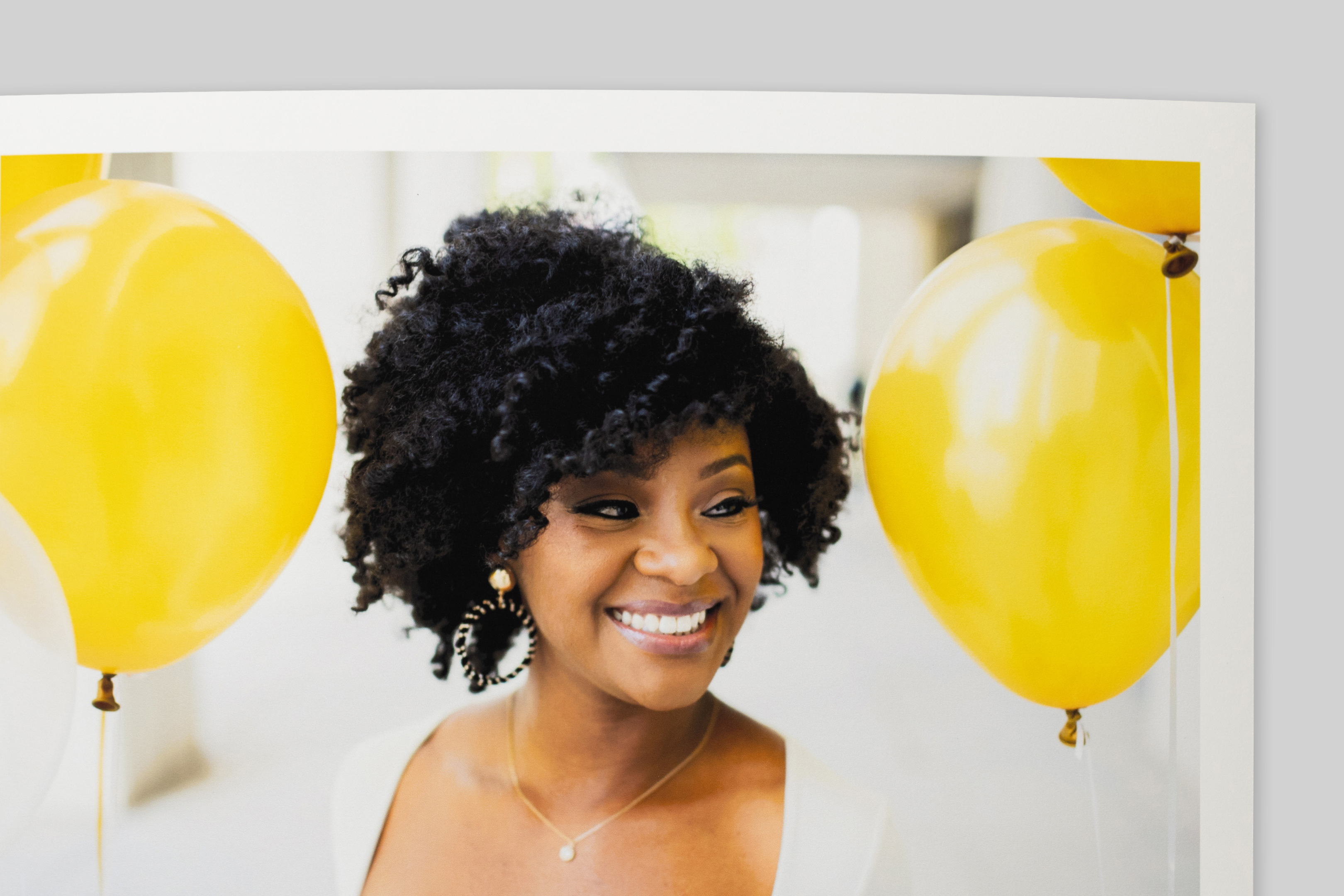 Corner of Smooth Matte Fine Art Print with border, featuring a portrait of a smiling woman with curly hair amid bright yellow balloons